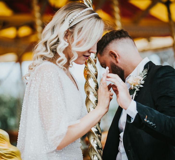 Bride in a sparkly wedding dress with curly hair and a double pearl headband holding hands with her groom on the carousel at Dreamland Margate 