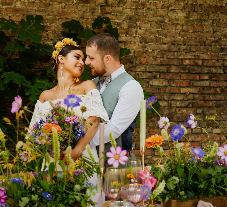 Groom embracing his boho bride in a strapless wedding dress and yellow headband standing behind a table full of colourful wildflowers and candles 