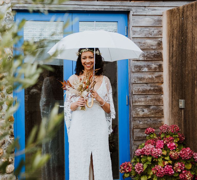 Bride stands outside blue door whilst holding white umbrella and carrying dried floral bouquet 