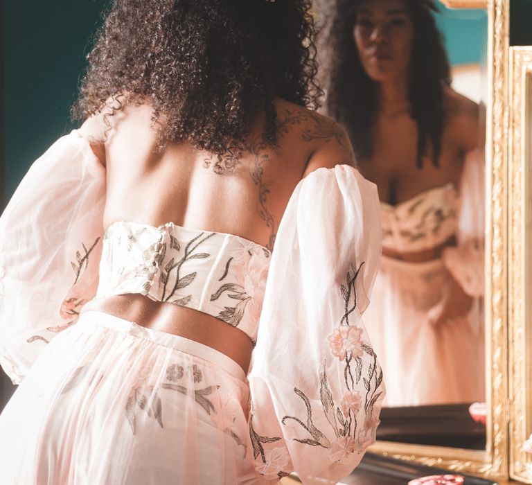 Black bride with long curly hair in blush pink bridal separates with appliqué detail looking in the mirror