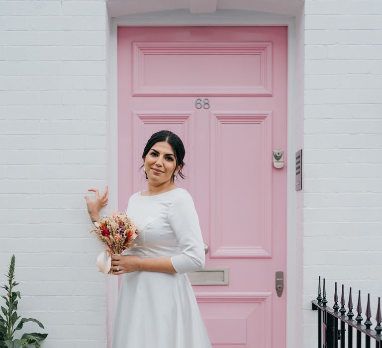 Bride stands in front of pastel pink door whilst holding dried floral bouquet and wearing PionDress wedding gown from Etsy