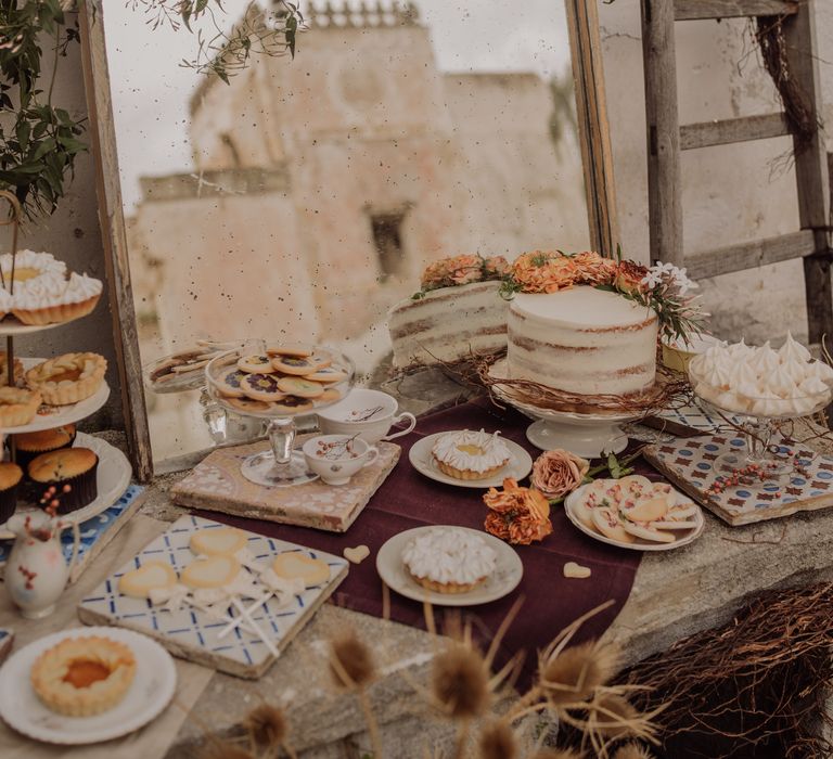 Buffet style dessert table on stone complete with rustic styled mirror surrounded by green hanging foliage