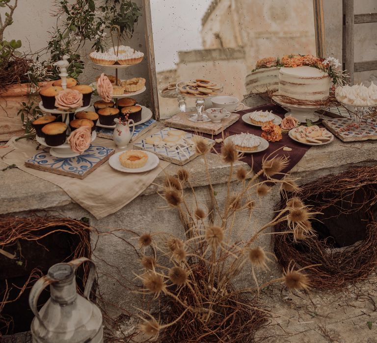 Buffet style dessert table set on stone with rustic style mirror leaning against wall and surrounded by dried floral arrangements