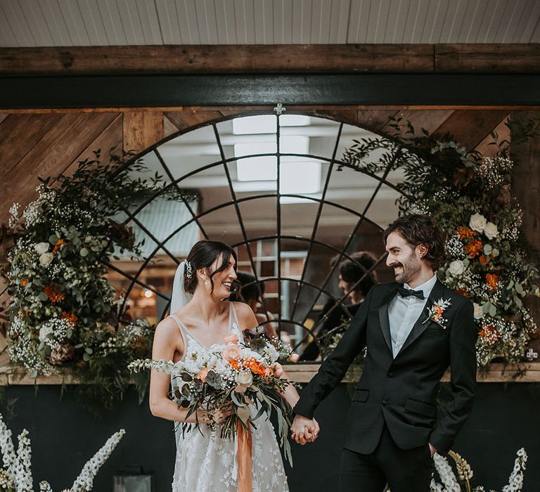 Bride in an appliqué wedding dress and groom in a tuxedo holding hands at the altar at new Lake District wedding venue, Ghyll Barn 