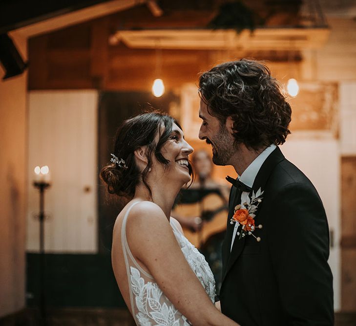 Groom in a black tuxedo with orange buttonhole smiling at his bride in a lace wedding dress with curly bridal up do 