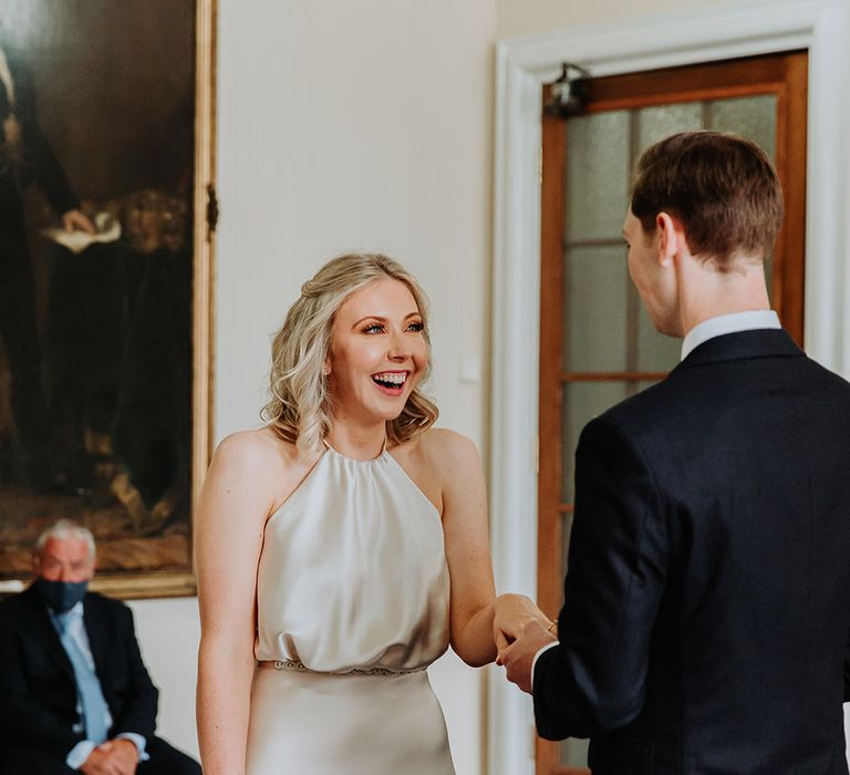 Smiley bride in a halterneck, champagne colour wedding dress holding hands with her husband during their registry office wedding 