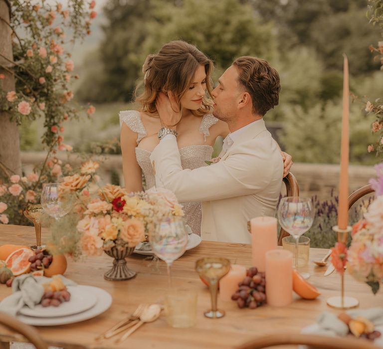 Groom in a stone coloured suit embracing his brides face at their outdoor wedding reception with orange candles, gold accents and blush flowers 