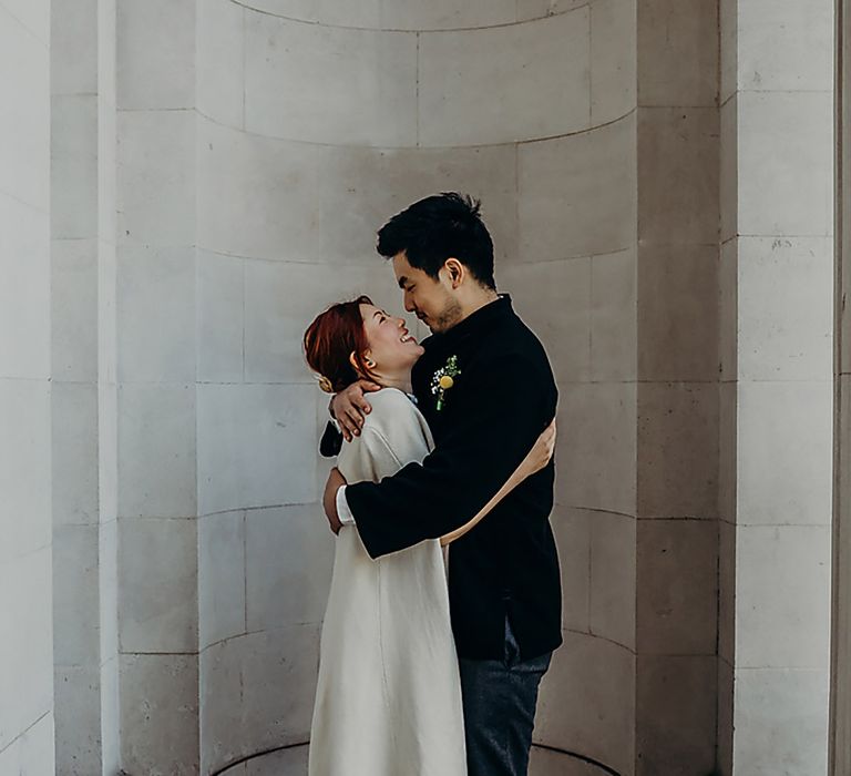 Groom in a velvet navy jacket embracing his bride in a cape coat outside Old Marylebone Town Hall 