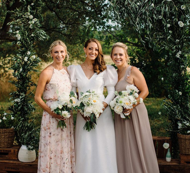 Bride stands with her bridesmaids who wear different style and coloured bridesmaids dresses whilst holding white floral bouquets