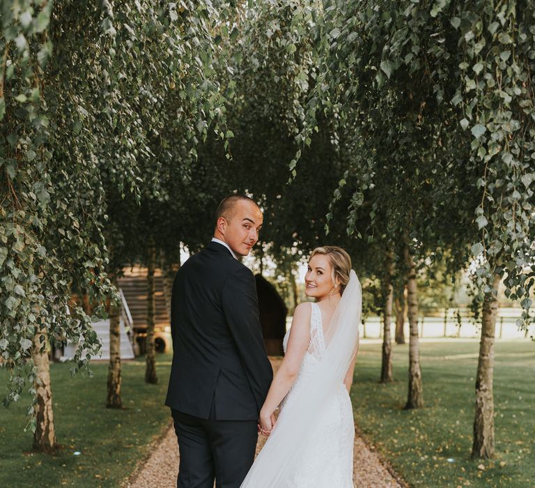 Bride in white lace Justin Alexander wedding dress and veil holds hands with groom in dark suit as they both look over their shoulders in the grounds at Tythe Barn wedding with barn wedding flowers