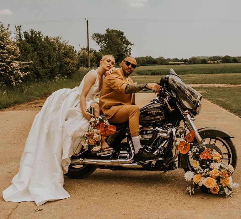 Stylish bride and groom in a Jesus Peiro wedding dress and mustard suit sitting on the back of a Harley Davidson Motorcycle 