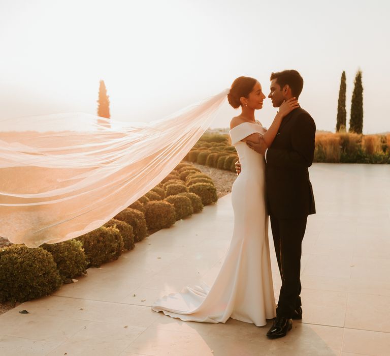 Bride & groom embrace outdoors as brides veil blows behind them during sunset in Greece | Hannah MacGregor Photo & Film