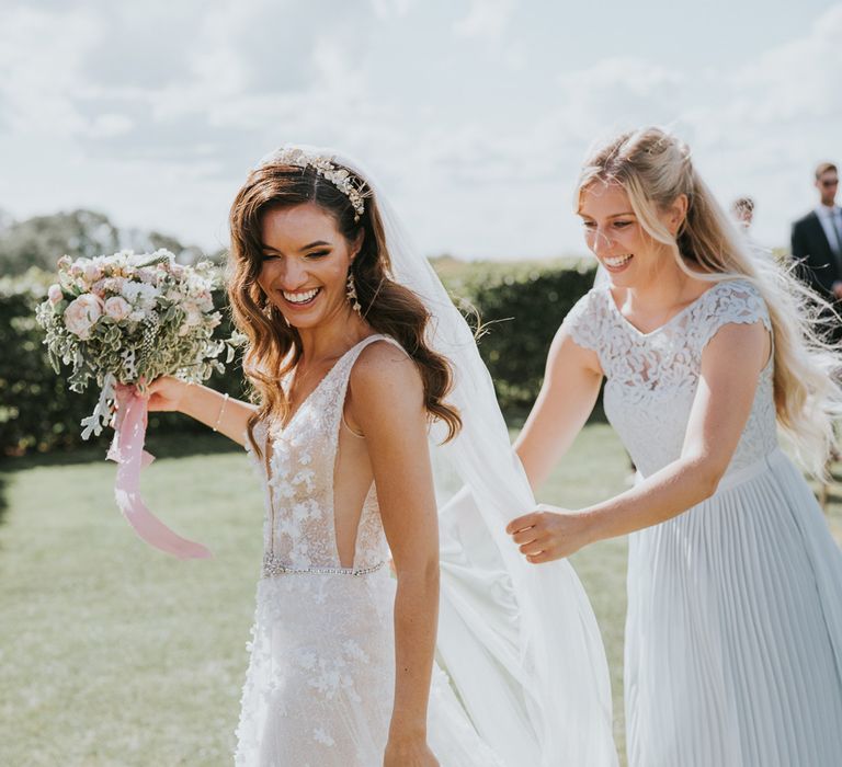 Bride in applique Berta wedding dress with headband and veil holding mixed bridal bouquet walks with wedding guests in blue dress adjusting her veil during summer wedding at Primrose Hill Farm