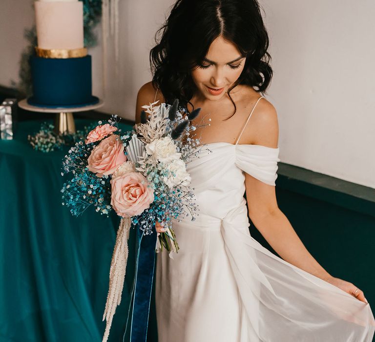 Bride with shoulder length wavy hair in a cold shoulder wedding dress holding her pink rose and teal gypsophila wedding bouquet tied with velvet ribbon 