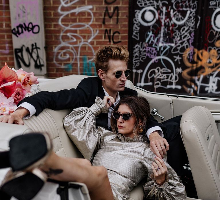 Edgy Bride wears tortoise shell sunglasses and sunflower earrings with groom wearing ray bans sitting in vintage car