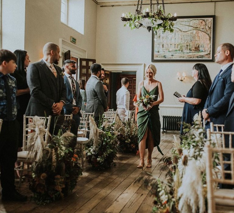 Bridesmaid walks down the aisle with her blonde hair tied in a low up-do and wearing a deep green bridesmaids gown