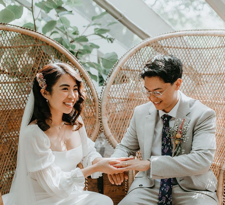 Bride & groom hold hands as they sit within wicker chairs 