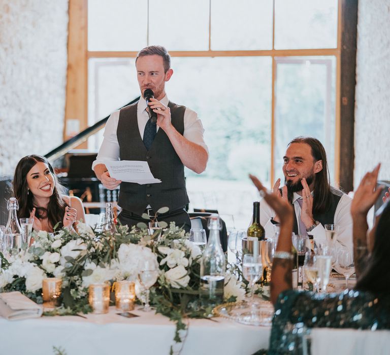 Groom in white shirt and grey waistcoat makes speech from round top table at wedding reception in Buckinghamshire