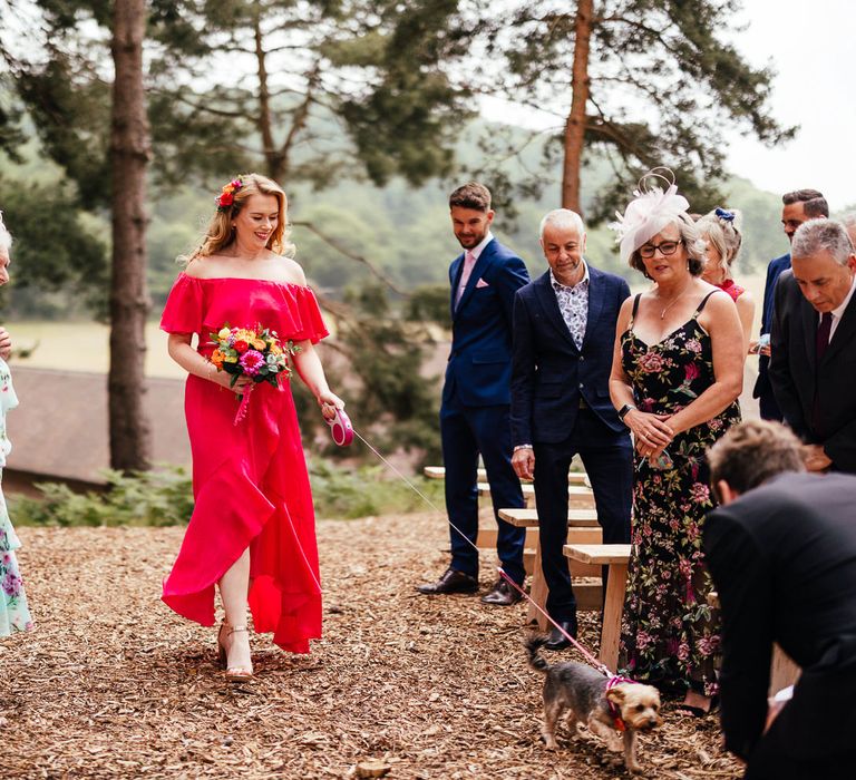 Bridesmaid in hot pink off the shoulder bridesmaid dress carrying colourful wedding bouquet walks dog down the aisle at woodland wedding ceremony 