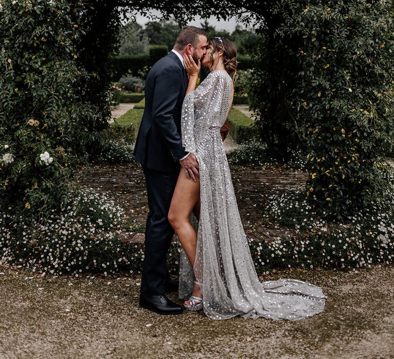 Bride in a silver sequin wedding dress and groom in a navy suit kissing in front of a greenery arch 