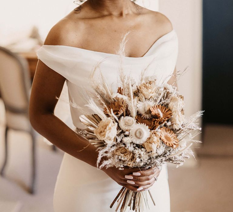 Bride wears off the shoulder wedding gown complete with dried floral bouquet