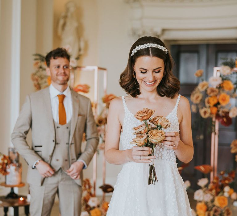 Bride with bobbed wavy brown hair in a white pearl headband holding a posy of orange roses 