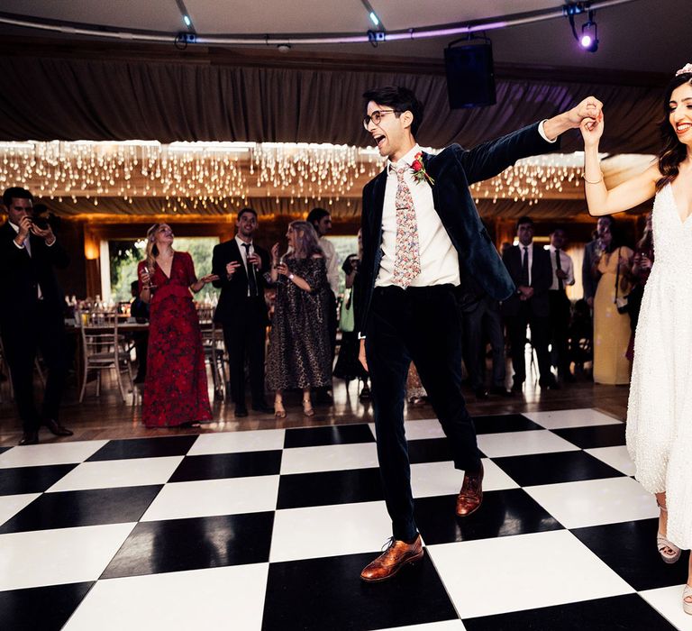 Groom in dark suit and floral tie leads bride in white ASOS midi dress onto the checkerboard dance floor at wedding