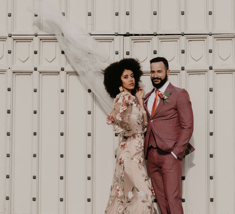 Black bride in a long sleeve pink embroidered wedding dress and veil with her groom in a dusky pink suit at Chiddingstone Castle 