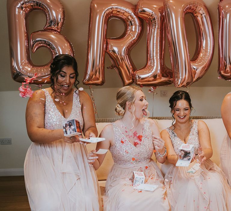 Bridesmaids in matching pale pink sparkly dresses opening gifts in front of rose gold balloons