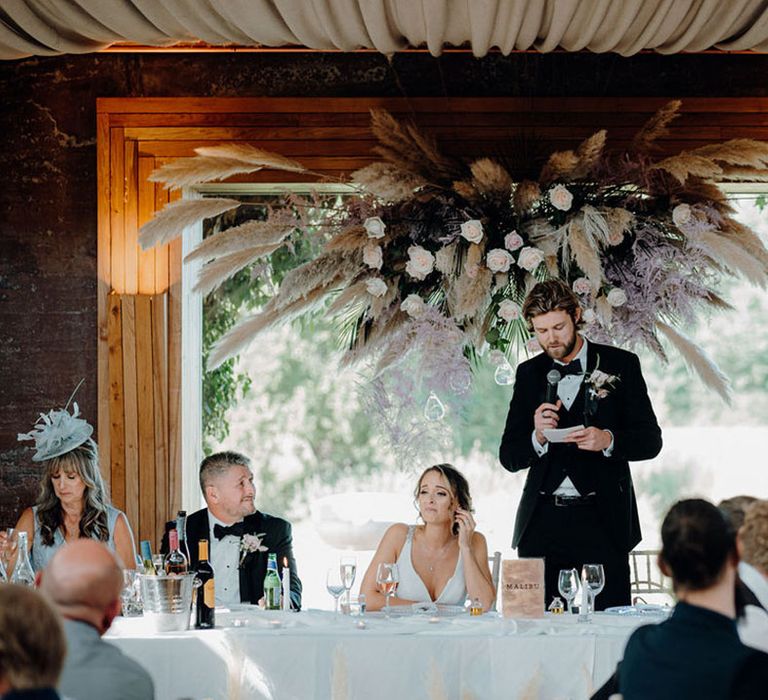 Groom in black tux and bride in mermaid fishtail wedding dress under pampas grass, foliage and rose flower installation 