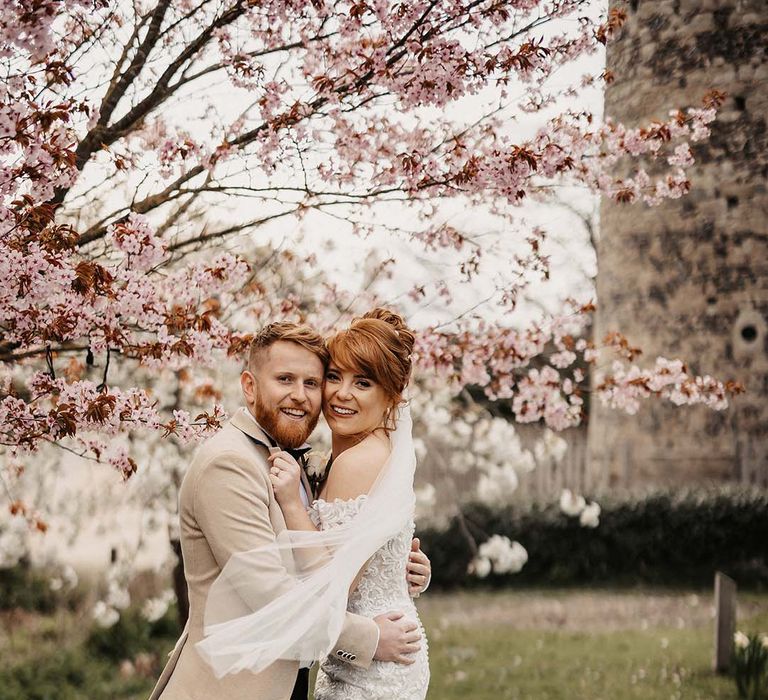 Bride and groom embrace under blossom trees for wedding in spring at rustic barn venue