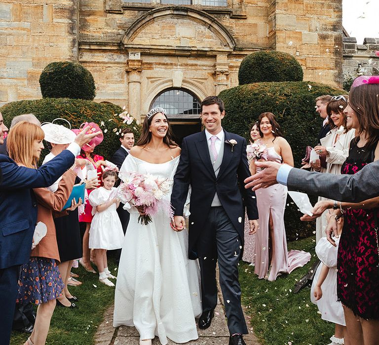 Wedding guests all wear shades of pink for marquee wedding as bride and groom walk through confetti 