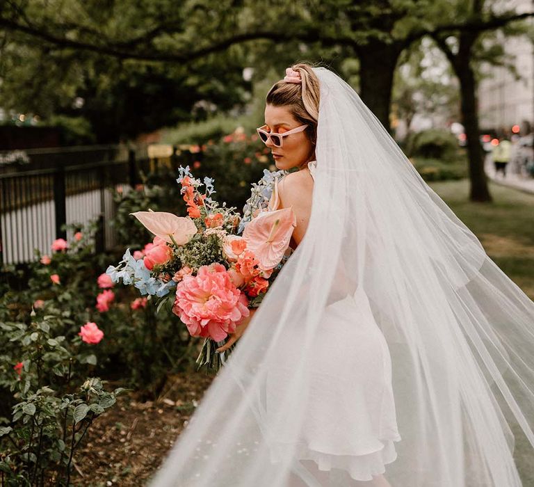 Bride in a long veil and sunglasses holding a coral pink and blue wedding bouquet with peony, anthuriums and delphiniums