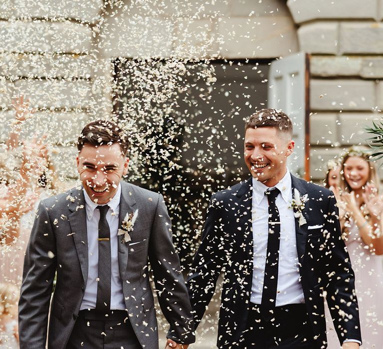 Grooms in grey and black suit with ties and matching gold tie clips have their confetti exit with dried flower buttonholes with white feather