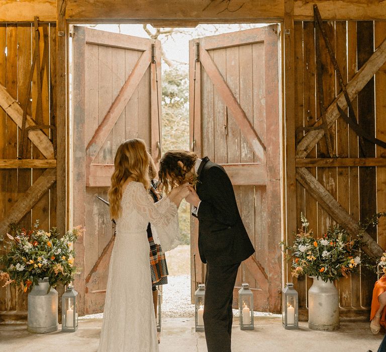 Groom in black tie kisses the bride's hands in lace wedding dress at altar with silver lanterns and candles