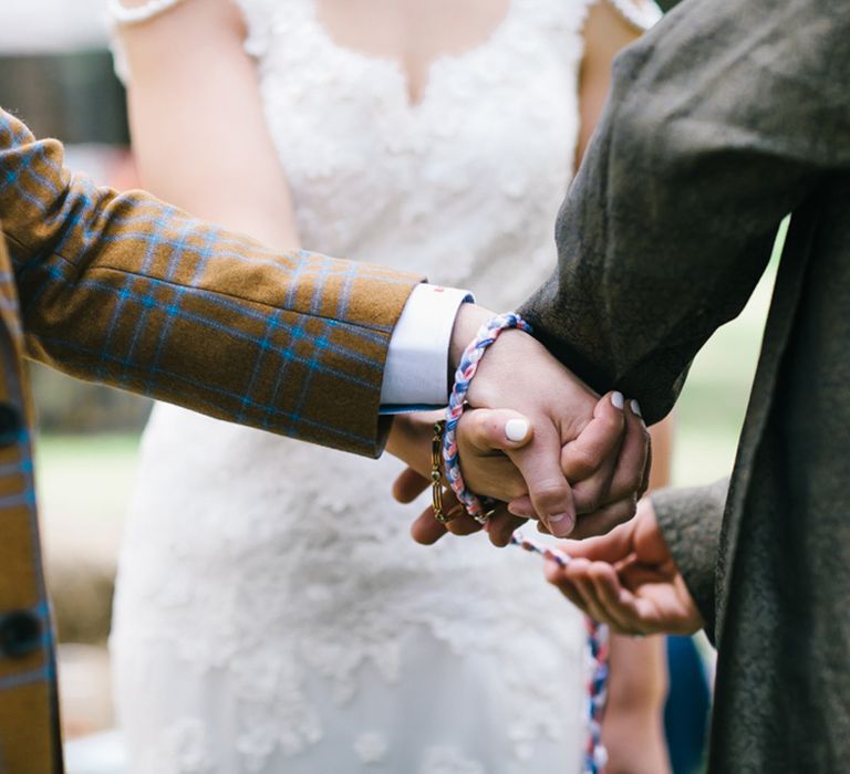 Outdoor boho wedding with groom in a check suit holding hands with his bride during the hand fasting ceremony