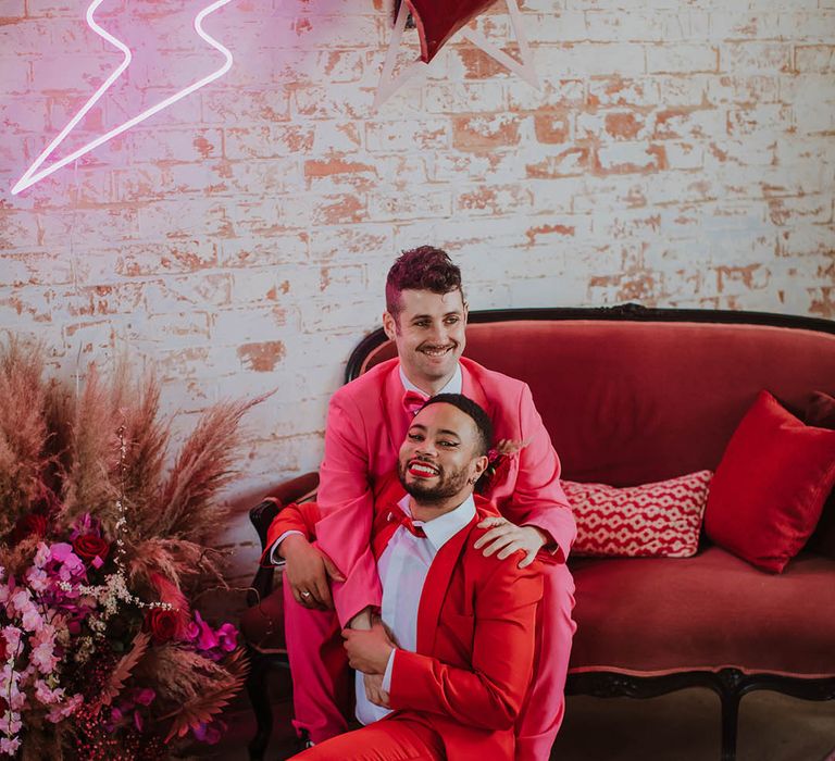 LGBTQI+ couple at Prestwold Hall Barn wedding sitting on a velvet couch surrounded by quirky neon sign and heart wedding decor 