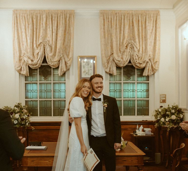 Bride in puff sleeve Story of My Dress wedding dress and long embroidered veil holds hands with groom in black tie after their registry office wedding |