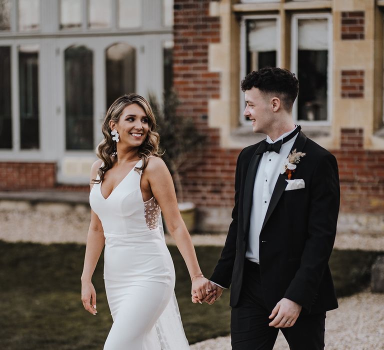 Bride in Made With Love Bridal wedding dress with mesh back with dot design and white flower earrings walks holding hands with groom in black tie