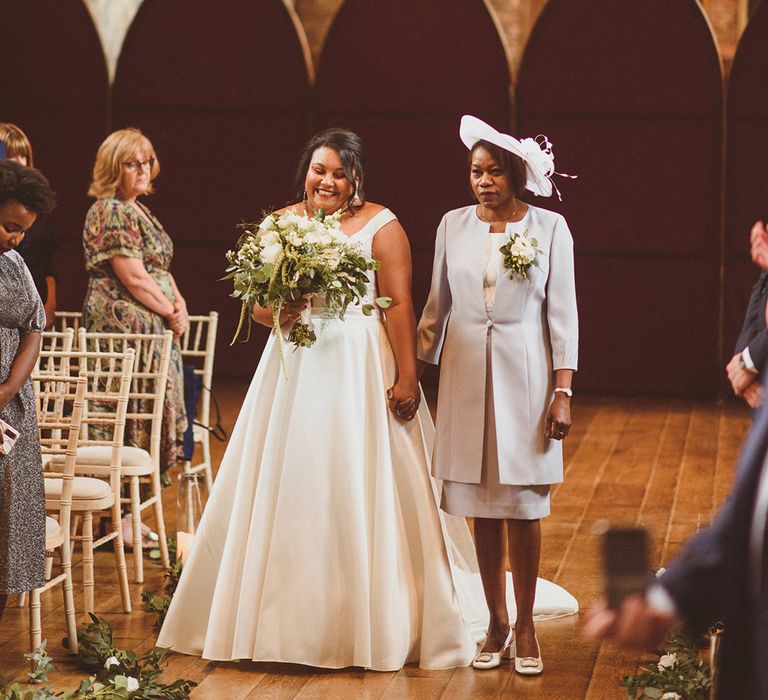 Mother of the bride wearing white flower buttonhole walks the bride in princess skirt wedding dress down the aisle