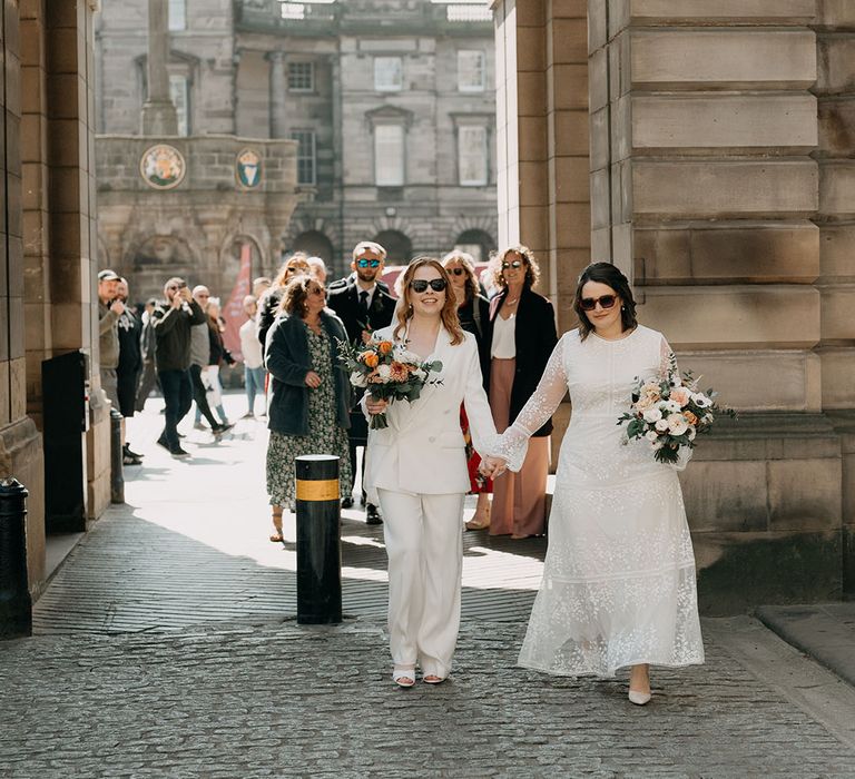 Two brides walk with one another whilst wearing sunglasses and carrying bouquets filled with white and orange florals 