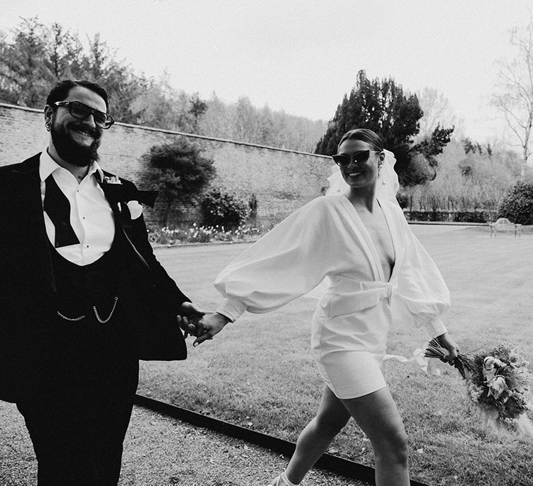 Groom in a horseshoe waistcoat with bow tie holding hands with his bride in a short wedding dress and sunglasses in the gardens at Garthmyl Hall