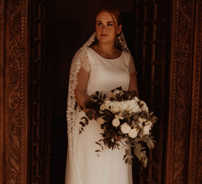 Bride wears round neck wedding gown with lace sleeves holding white rose floral bouquet and wearing floor length veil