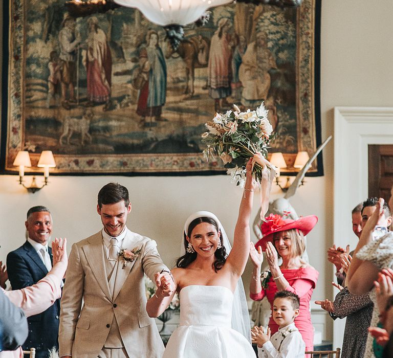 Bride in strapless pleated and brocade Jesus Peiro wedding dress and headband walking back down the aisle with the groom in a neutral wedding suit