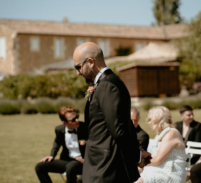 Groom wears black tie suit with pale pink floral buttonhole and sunglasses whilst waiting for his bride 