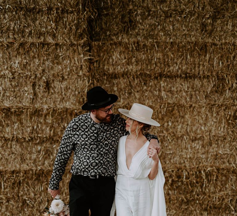 Bride and groom in cool hats for their with the bride wearing a jumpsuit with a plain cape 