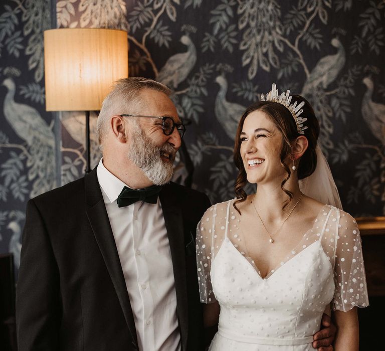 Father of the bride in black tie smiles at the bride wearing an epic crystal crown and a dotted wedding dress 