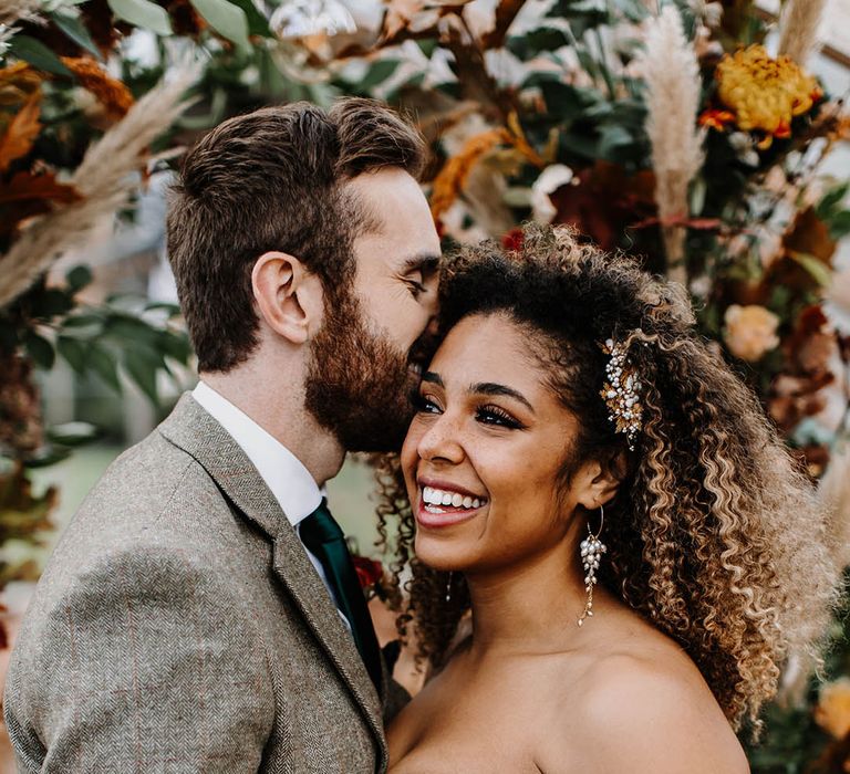 Black bride with a gold and pearl hair clip and matching earrings with an off the shoulder structured bodice wedding dress and groom in grey tweed suit 