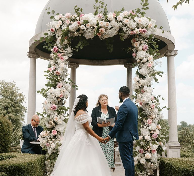 Bride wears off the shoulder wedding dress and stands beneath wedding dome complete with floral archway 