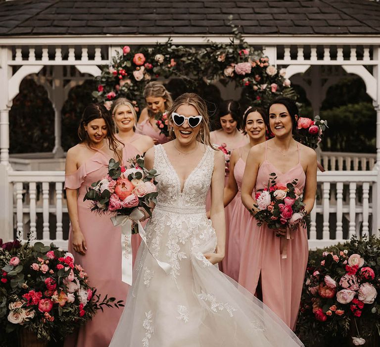 Bride in retro cat eye sunglasses in lace wedding dresses with bridesmaids in pink dresses in front of the pink flower arch